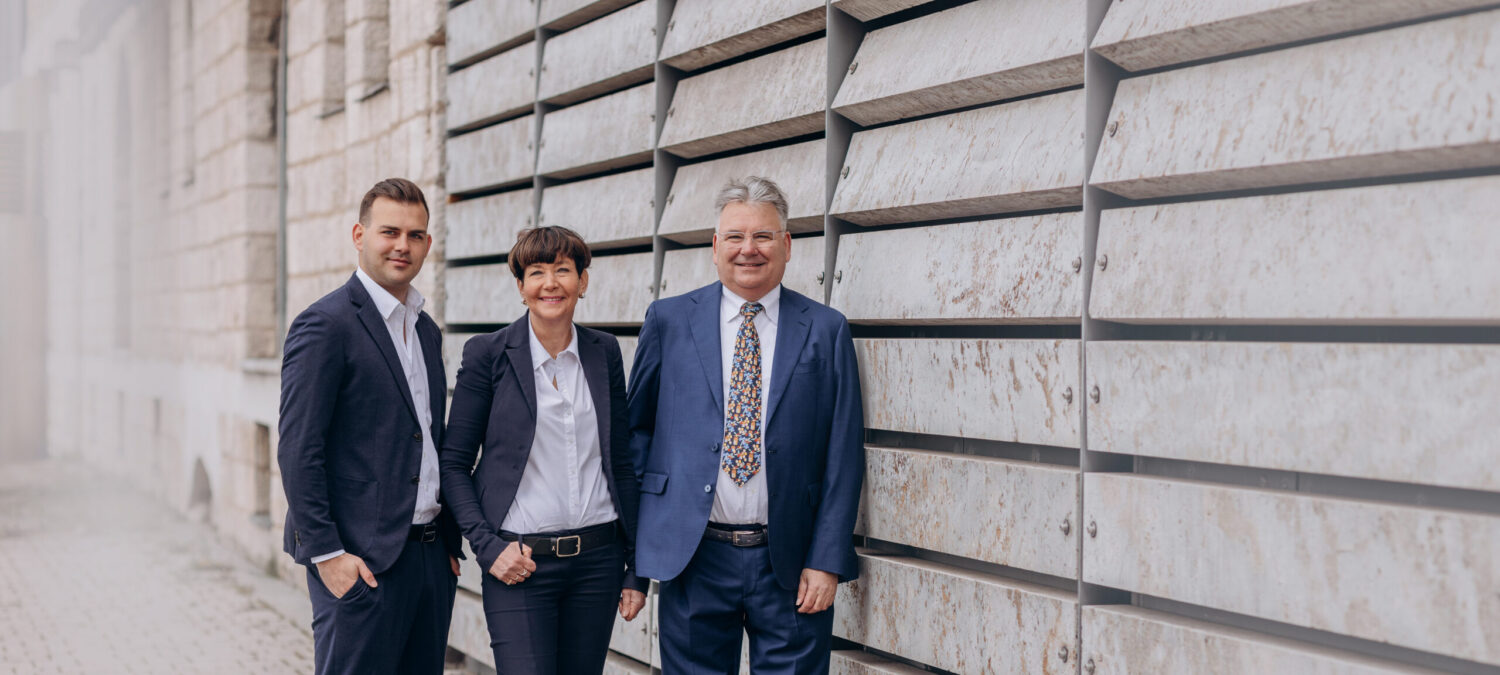 Zwei Männer und eine Frau in dunkler Businesskleidung stehen in die Kamera lächelnd nebeneinander auf einem gepflasterten Weg vor einer großflächigen, waagerecht strukturierten Metallfassade mit Natursteinoptik.