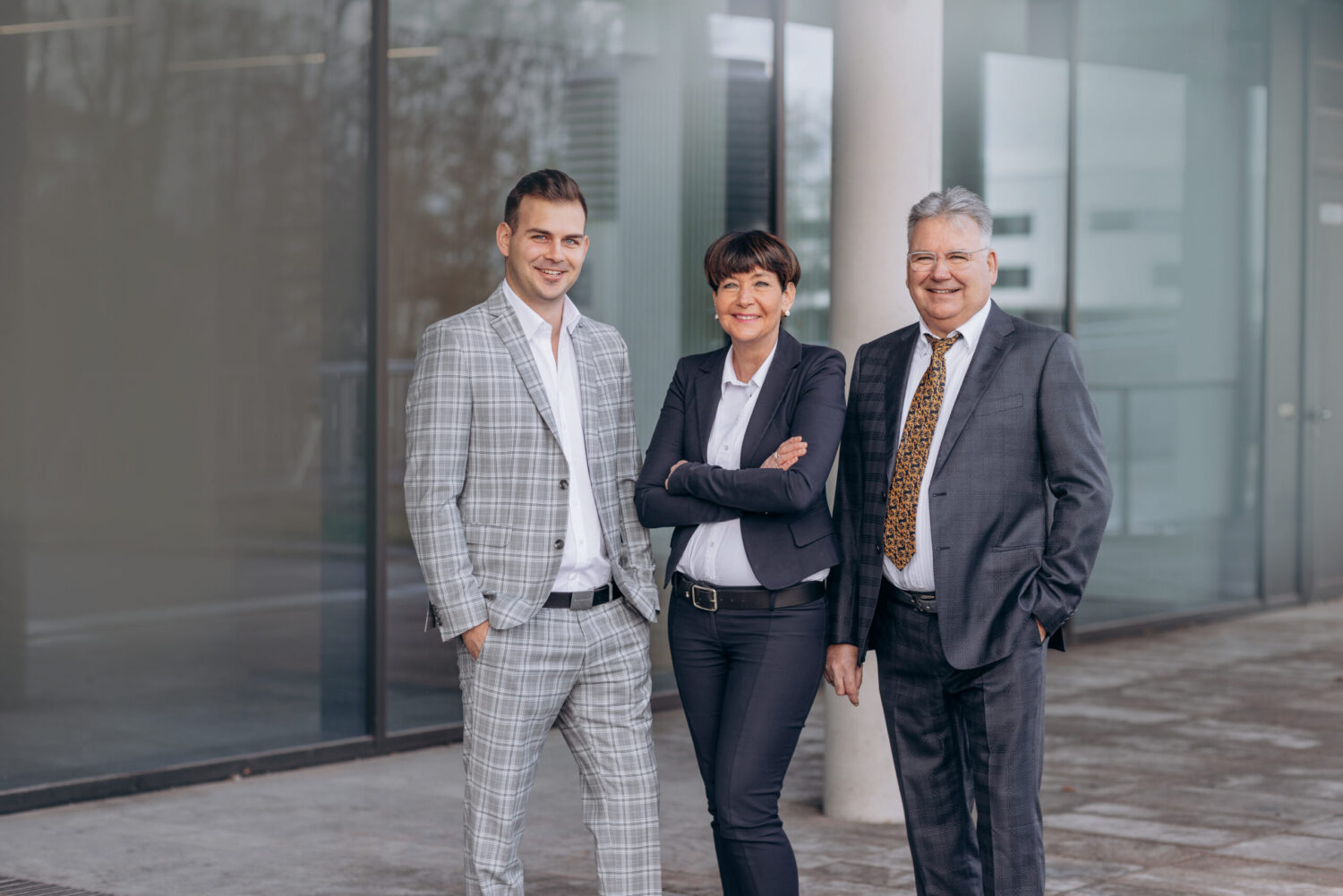 Zwei Männer und eine Frau stehen auf grauem Pflaster vor einem modernen Gebäude mit Fensterfront.