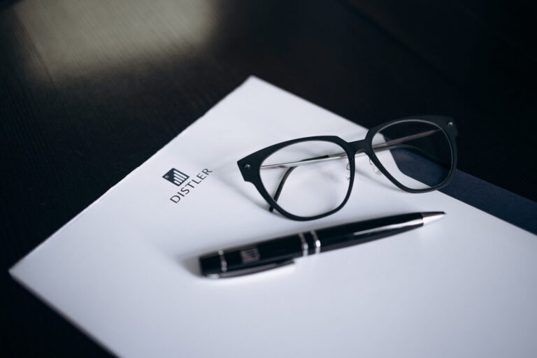 Schwarze Brille und schwarzer Kugelschreiber mit silbernen Details liegen auf einem weißen Schreibblock mit DISTLER-Logo, Hintergrund ist eine dunkle Tischoberfläche.