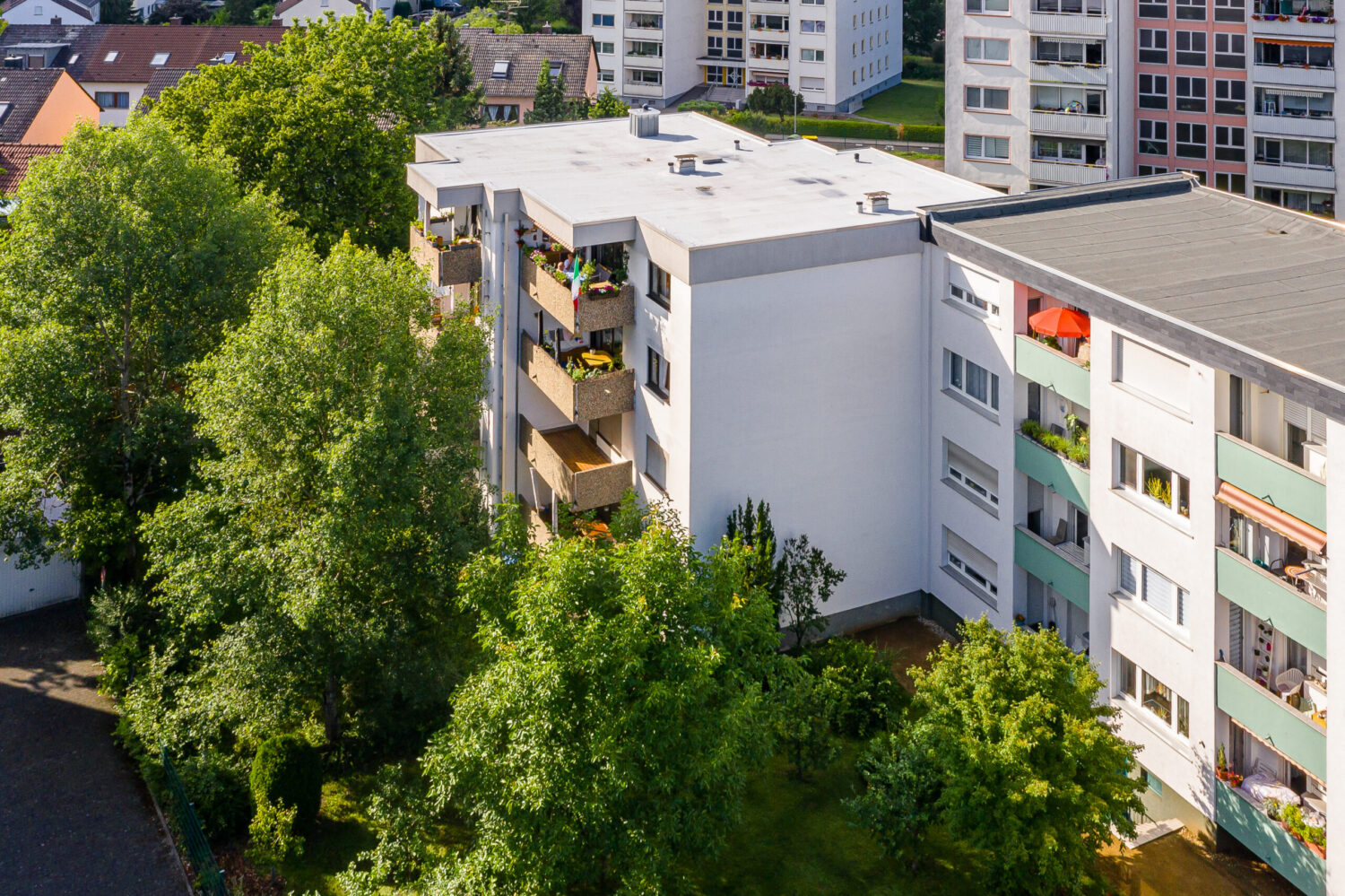 Fünfgeschossiges Wohnhaus mit Flachdach, weißer Putzfassade und mehreren bepflanzten Balkonen, umgeben von Bäumen; im Hintergrund weitere Wohngebäude sichtbar.