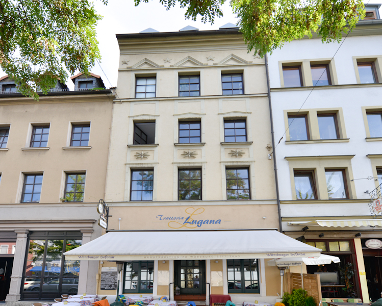 Viergeschossiges Gebäude mit heller Fassade, symmetrischer Fensteranordnung und Markise im Erdgeschoss, darunter ein Restaurant mit Außengastronomie und weißen Tischen.