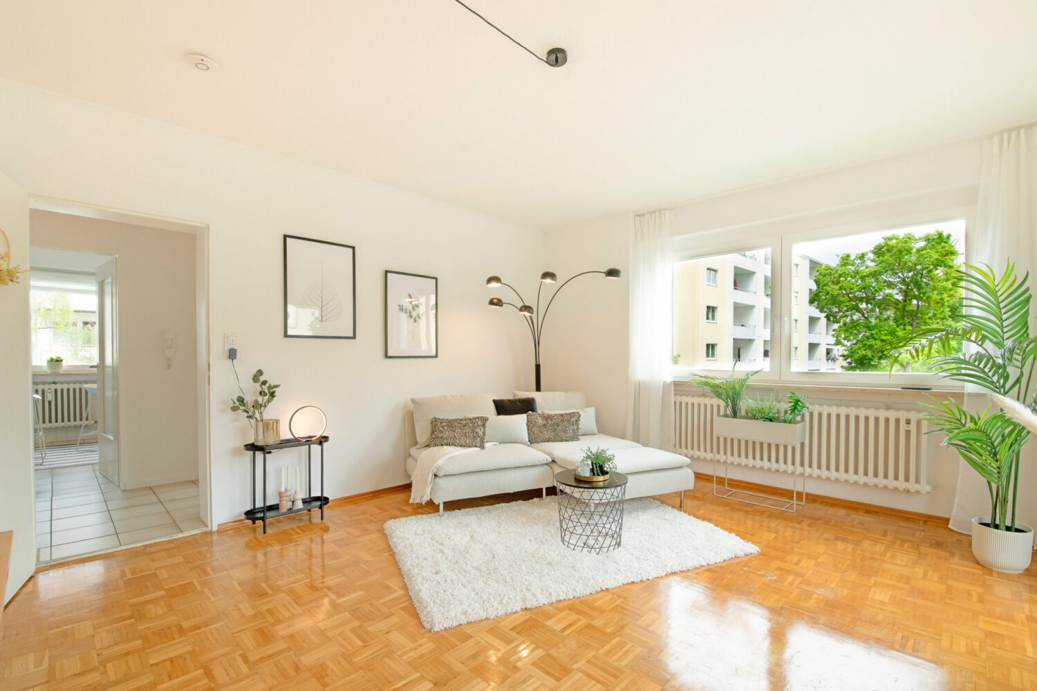 Modern eingerichtetes Wohnzimmer mit glänzendem Parkettboden, weißem Sofa, Teppich, runder Couchtisch, Bogenlampe, Pflanzen am Fensterbrett und Blick auf Nachbargebäude durch das Fenster.