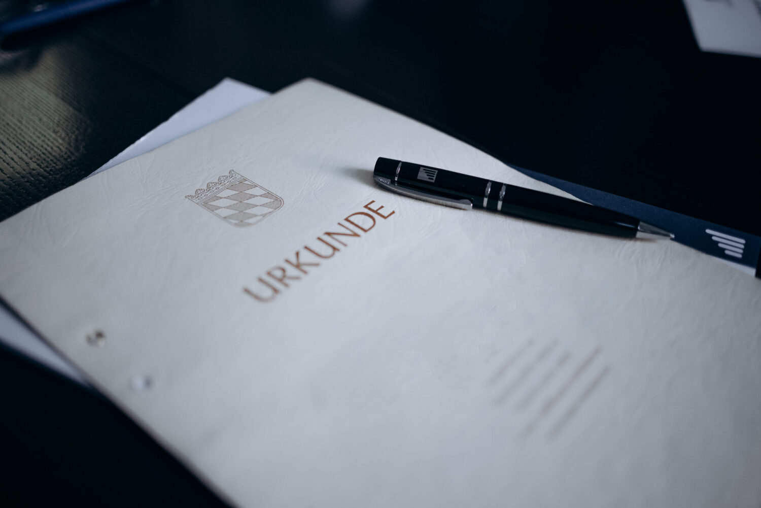 Beige Urkunde mit bayerischem Rautenmuster und der Aufschrift „URKUNDE“ liegt auf einem Stapel Papier; darauf ein schwarzer Kugelschreiber mit silbernem Clip und DISTLER-Aufdruck, Hintergrund ist eine dunkle Tischoberfläche.