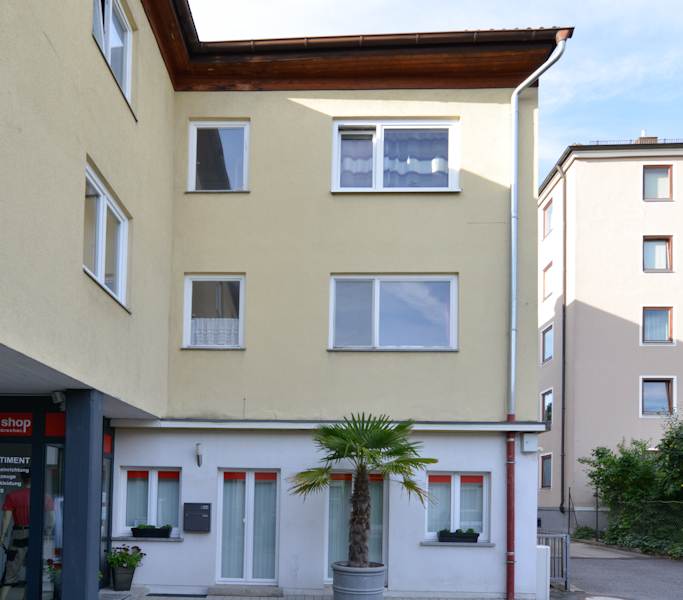 Dreigeschossiges Wohnhaus mit gelber Fassade, weißen Fenstern, kleinem Innenhofbereich und zwei Kübelpalmen vor dem Eingang im Erdgeschoss.
