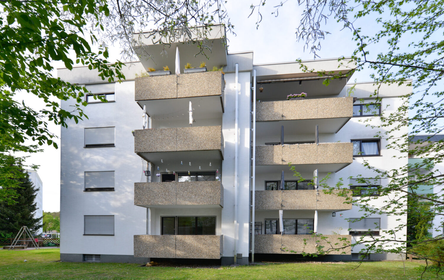 Mehrgeschossiges Wohngebäude mit außenliegenden Betonbalkonen, weißer Fassade, teils verdeckt durch Bäume im Vordergrund.
