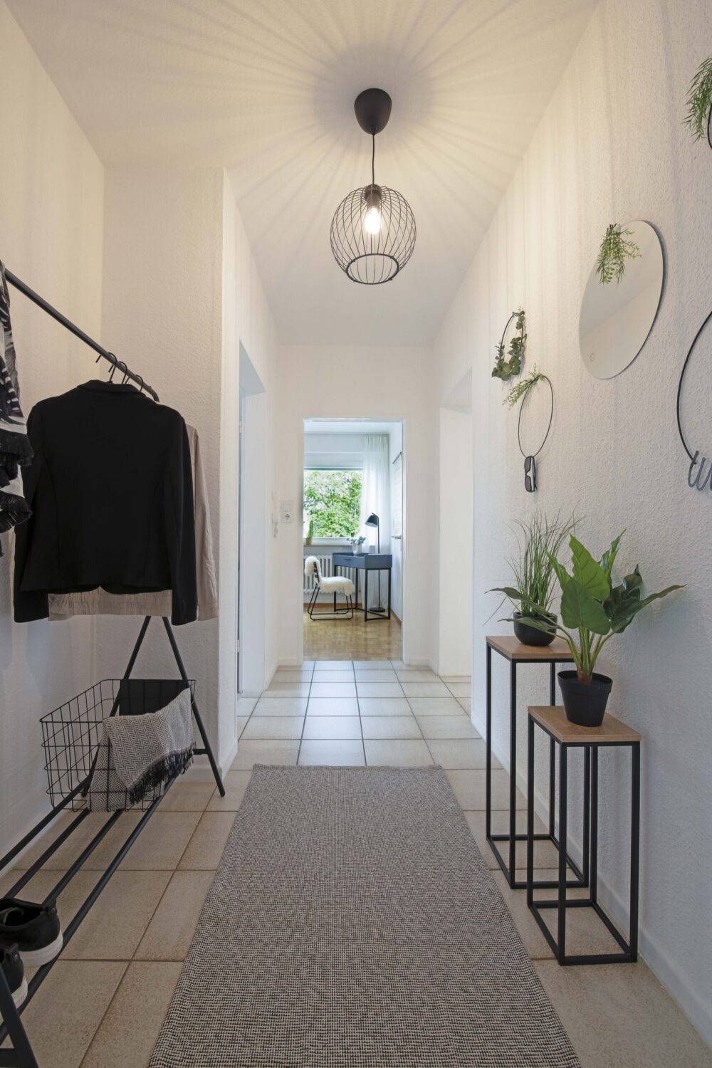 Langer Flur mit Fliesenboden, hellen Wänden, Pflanzen und runden Spiegeln an der rechten Wand, im Hintergrund ein Wohnraum mit Tageslicht.