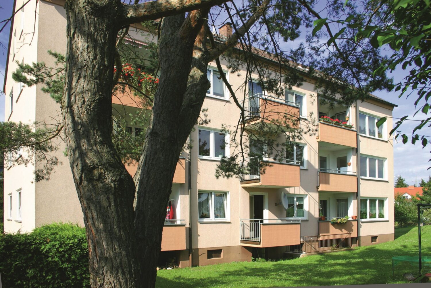 Dreigeschossiges Mehrfamilienhaus mit Erker, Balkonen und hellgelber Fassade, teils von Baumstämmen verdeckt.