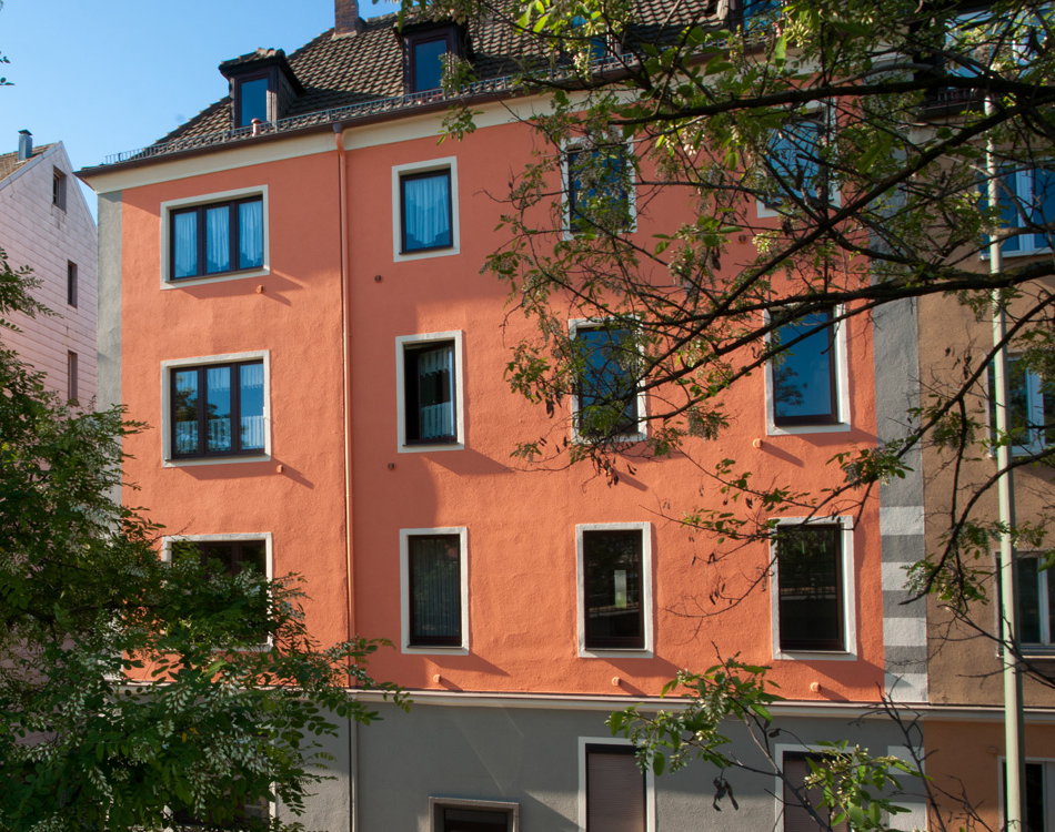 Viergeschossiges Wohnhaus mit orangefarbener Putzfassade, weißen Fensterrahmen und dunklem Dach, teils verdeckt durch Laubbäume im Vordergrund.