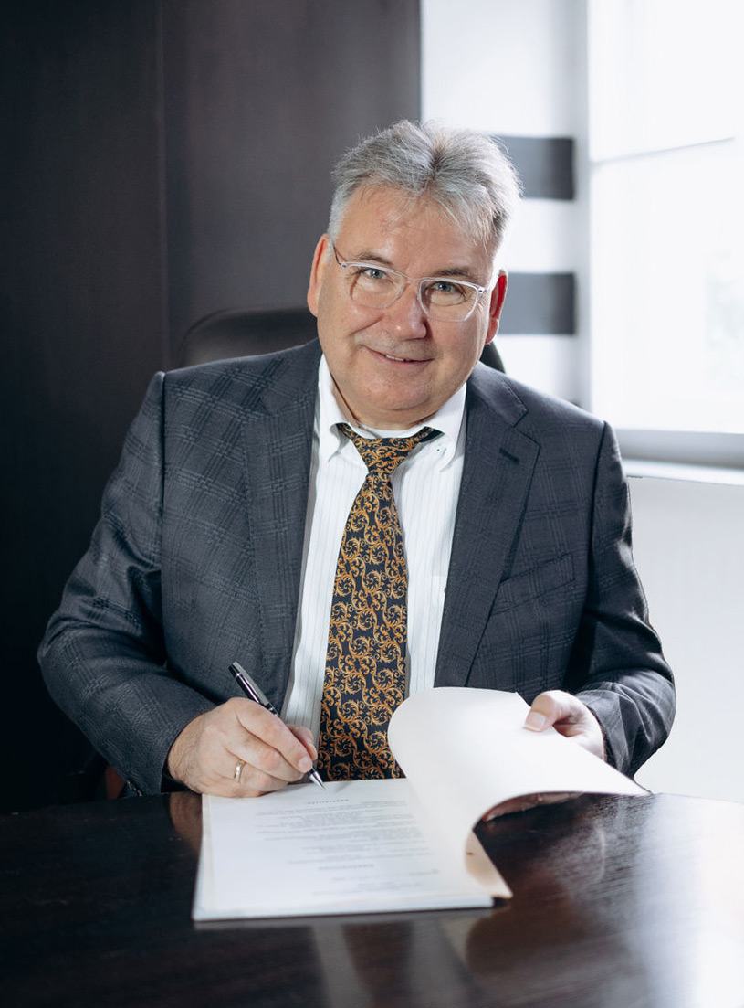 Mann mit grauen Haaren, Brille, gestreiftem Hemd, kariertem Sakko und gemusterter Krawatte sitzt an einem dunklen Tisch, hält einen Stift und ein geöffnetes Dokument.