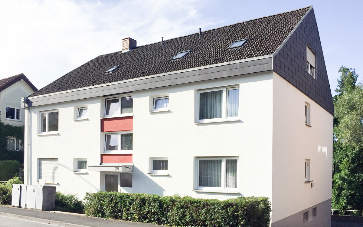 Dreigeschossiges Mehrfamilienhaus mit weißer Fassade, roten Akzenten, Satteldach und kleinen Fenstern, davor ein schmaler Vorgarten mit Hecke.