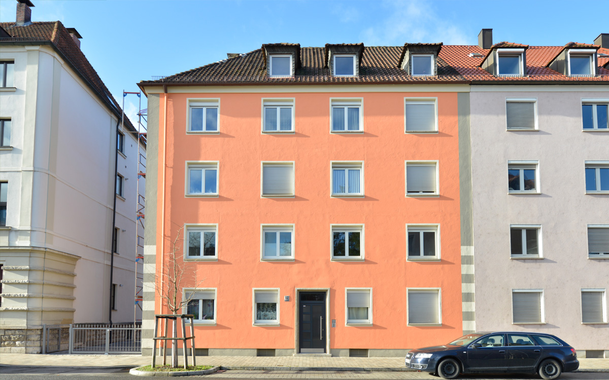 Fünfgeschossiges Wohnhaus mit orangefarbener Putzfassade, weißen Fensterrahmen, dunklem Satteldach und geparktem Auto im Vordergrund.