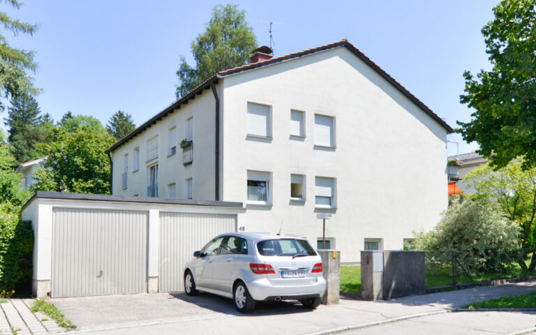 Rückansicht eines gepflegten Mehrfamilienhauses mit weißer Fassade, Garage, Parkplatz und Grünanlage.