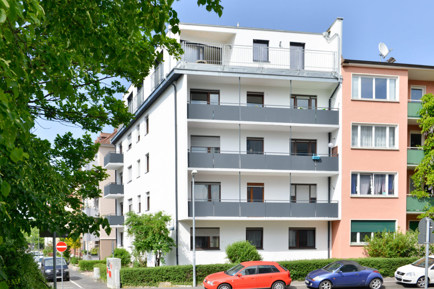Fünfgeschossiges weißes Wohngebäude mit durchgehenden Balkonen, Flachdach, grauen Brüstungen und parkenden Autos im Vordergrund.