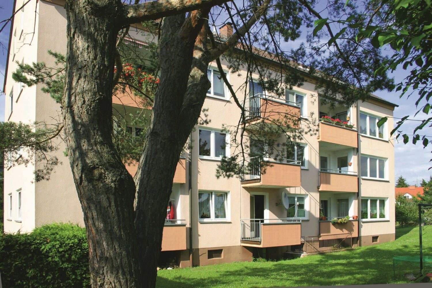 Dreigeschossiges Mehrfamilienhaus mit Erker, Balkonen und hellgelber Fassade, teils von Baumstämmen verdeckt.